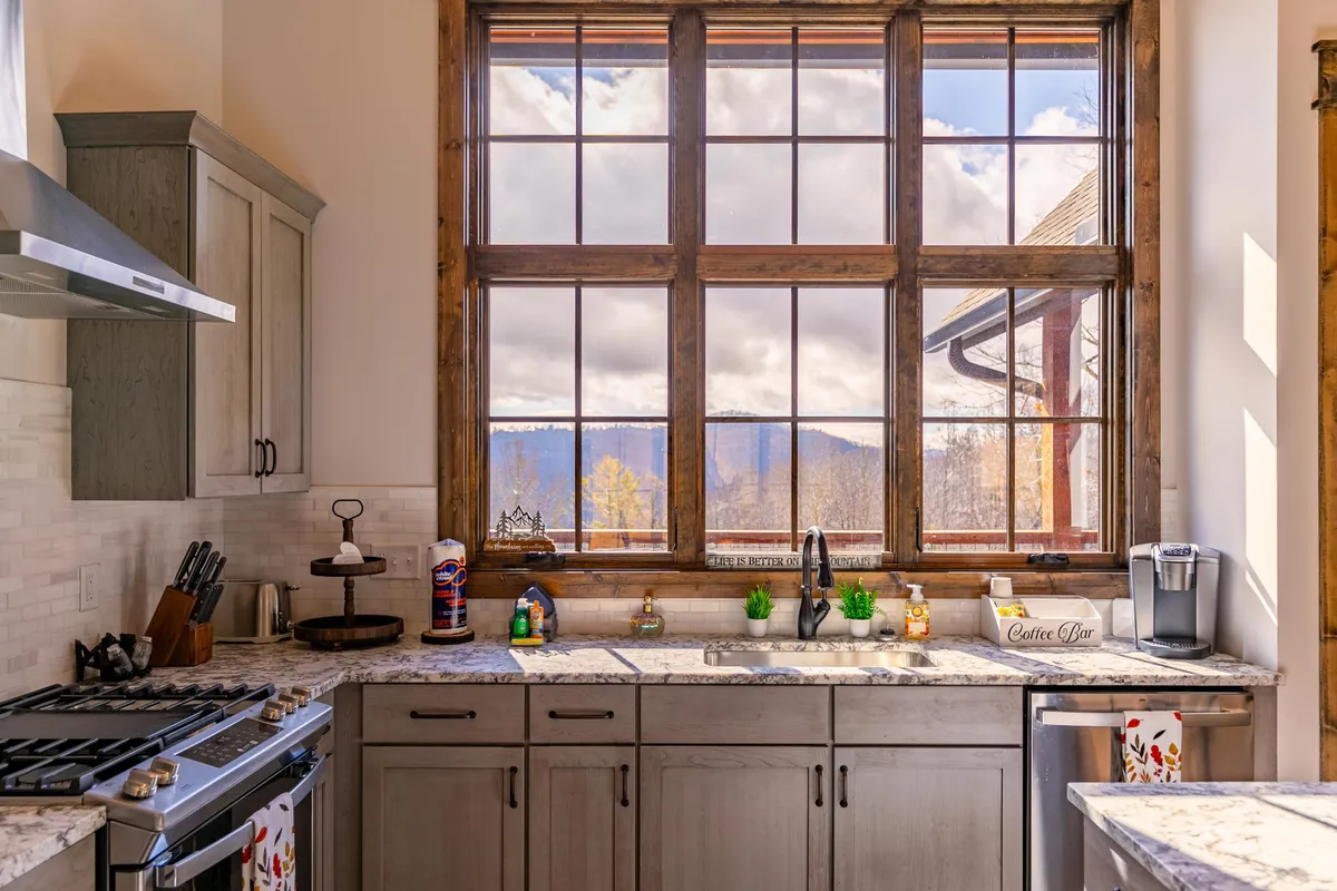 A cheerful kitchen space featuring a large window framing stunning mountain views. The countertops are adorned with decorative items, and cooking appliances are arranged neatly, creating a welcoming area for preparing coffee or meals. Natural light fills the room, enhancing the cozy appeal and emphasizing the scenic Blue Ridge backdrop.