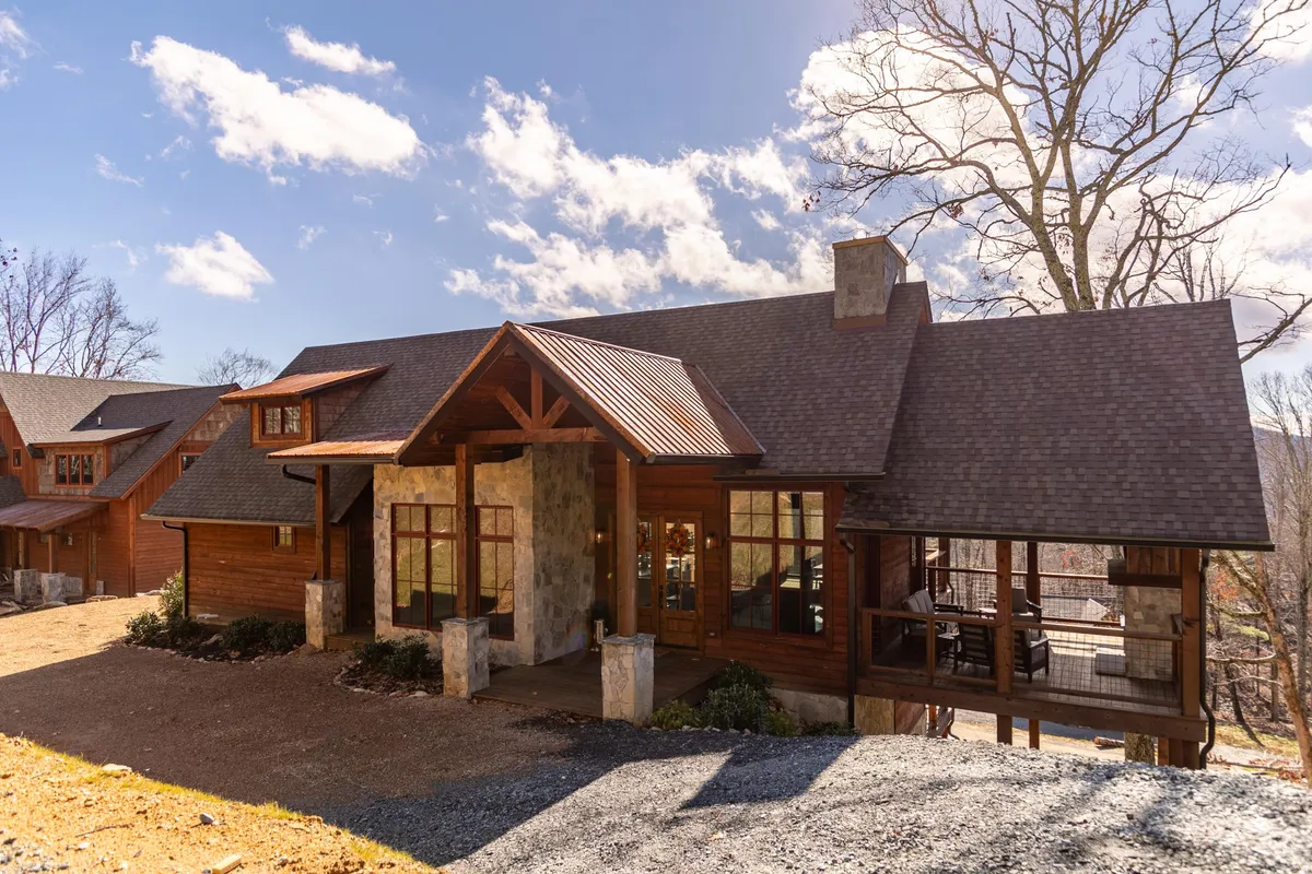 The image showcases the inviting exterior of Hucklebeary Cabin, emphasizing its charming wooden front porch, impressive stone facade, expansive windows, and the distinctive roofline. The welcoming porch hints at relaxing mornings with coffee, while the natural surroundings suggest a tranquil mountain getaway perfect for families or groups.
