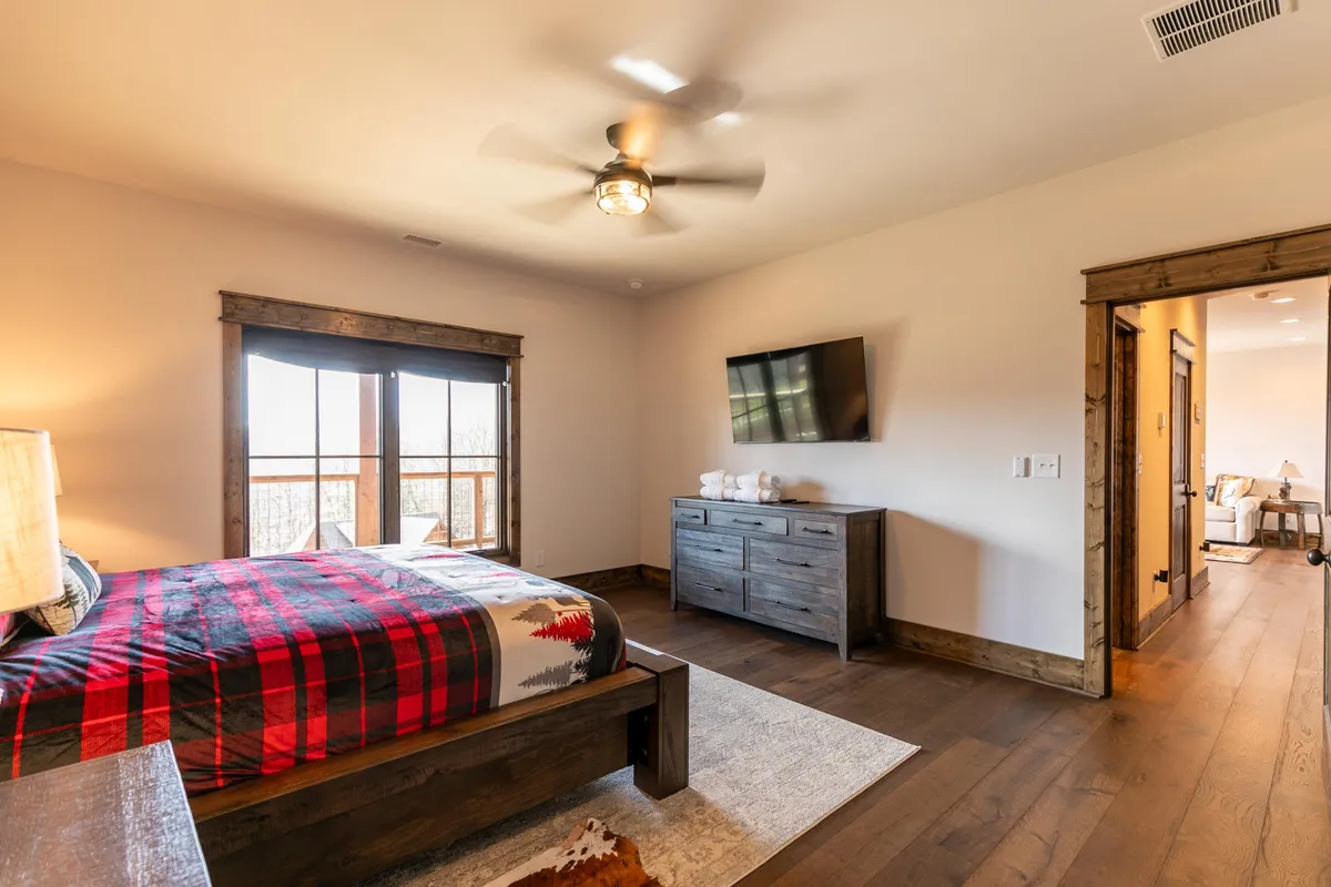 A cozy bedroom with rustic plaid bedding, wooden dresser, large windows framing mountain views, and a wall-mounted TV, offering a comfortable and inviting retreat for relaxation.