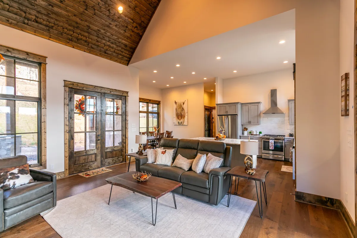 This image offers another perspective of the open-plan layout, showing the connection between the living area (sofa arrangement), kitchen, and large windows that let in natural light. The warm wood tones and well-appointed kitchen underscore the inviting, communal atmosphere.