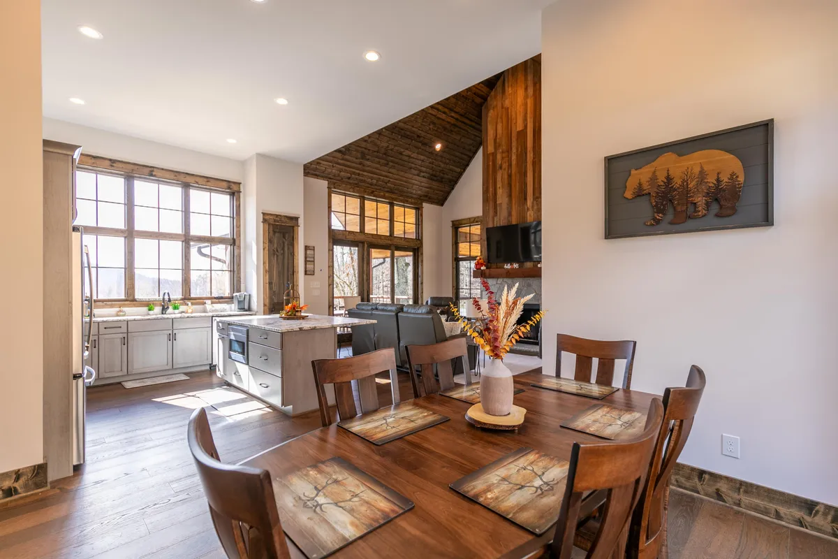 This image presents the kitchen and dining area from a complementary angle, focusing on the spacious dining table, the kitchen island, large windows, and a decorative wooden accent wall. A touch of personality comes through with the bear artwork, enhancing the homey, mountain vibe.