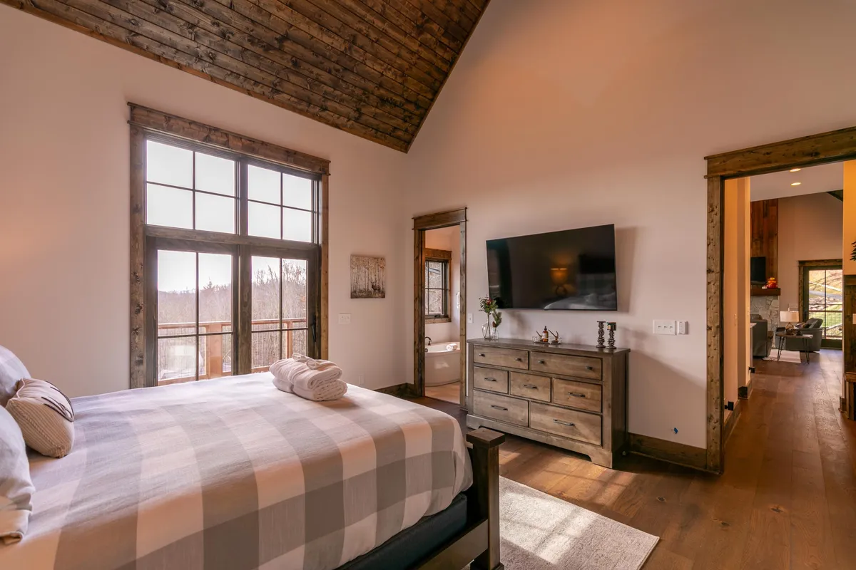 A serene bedroom featuring plaid bedding, wooden furniture, and large windows that let in ample natural light. The inviting bed and view suggest a tranquil retreat after a day of mountain adventures, offering both comfort and a visual connection to the outdoors.