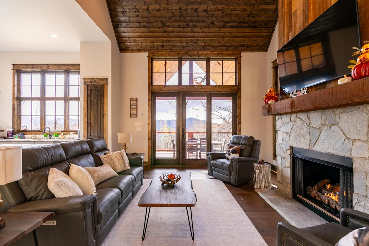 A welcoming living area featuring a striking stone fireplace, comfortable seating arranged for socializing, and expansive windows that reveal stunning mountain views. The warm tones and abundant natural light make it an inviting place to gather, relax, or watch the sunset together.
