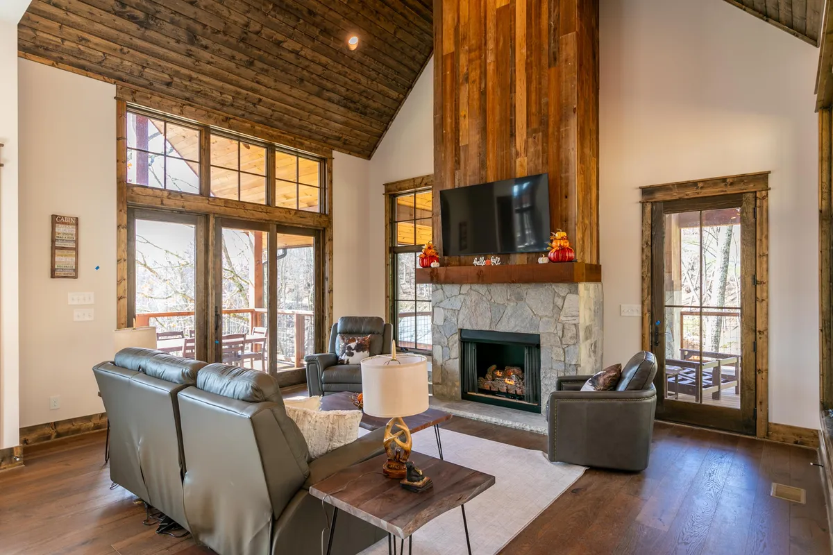 A spacious living area highlighted by a wooden fireplace, large windows offering picturesque views, and plush seating for group gatherings. The room is perfect for relaxing by the fire, watching movies, or enjoying nature through the windows.